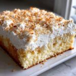 Close-up of a square slice of coconut poke cake topped with thick white frosting and toasted coconut flakes.