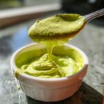 A spoonful of thick, bright green avocado sauce dripping back into a small white bowl.
