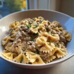 A close-up of a white bowl filled with creamy beef pasta made using farfalle (bowtie) noodles, garnished with parsley.