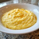 Close-up of a white bowl filled with creamy, bright yellow cheese grits, showing melted butter on top.