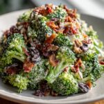 A close-up, appetizing shot of creamy broccoli salad mixed with bacon bits, dried cranberries, and nuts.