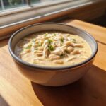 A close-up of a bowl of creamy white chicken chili, featuring shredded chicken, white beans, and green onion garnish.