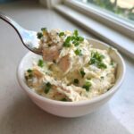 A spoonful of creamy clam dip topped with fresh chives being lifted from a white bowl.