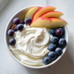 A white bowl filled with creamy fruit dip, topped with fresh blueberries, grapes, and sliced red apples.