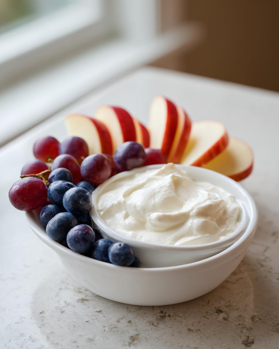 A bowl of creamy white fruit dip surrounded by fresh blueberries, red grapes, and sliced red apples.