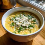 A close-up of a creamy, yellow Mexican street corn soup topped generously with crumbled white cheese and fresh cilantro.