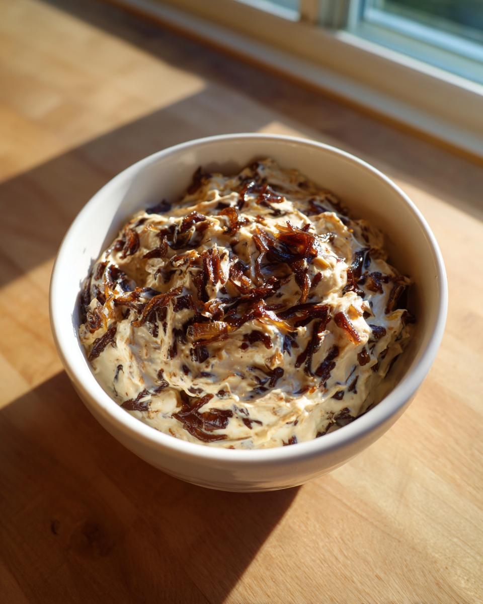 A white bowl filled with creamy onion dip topped generously with dark, caramelized onions, sitting on a wooden surface.