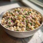 Close-up of a creamy Tuna Pasta Salad mixed with elbow macaroni, green peas, and red onion in a light-colored bowl.