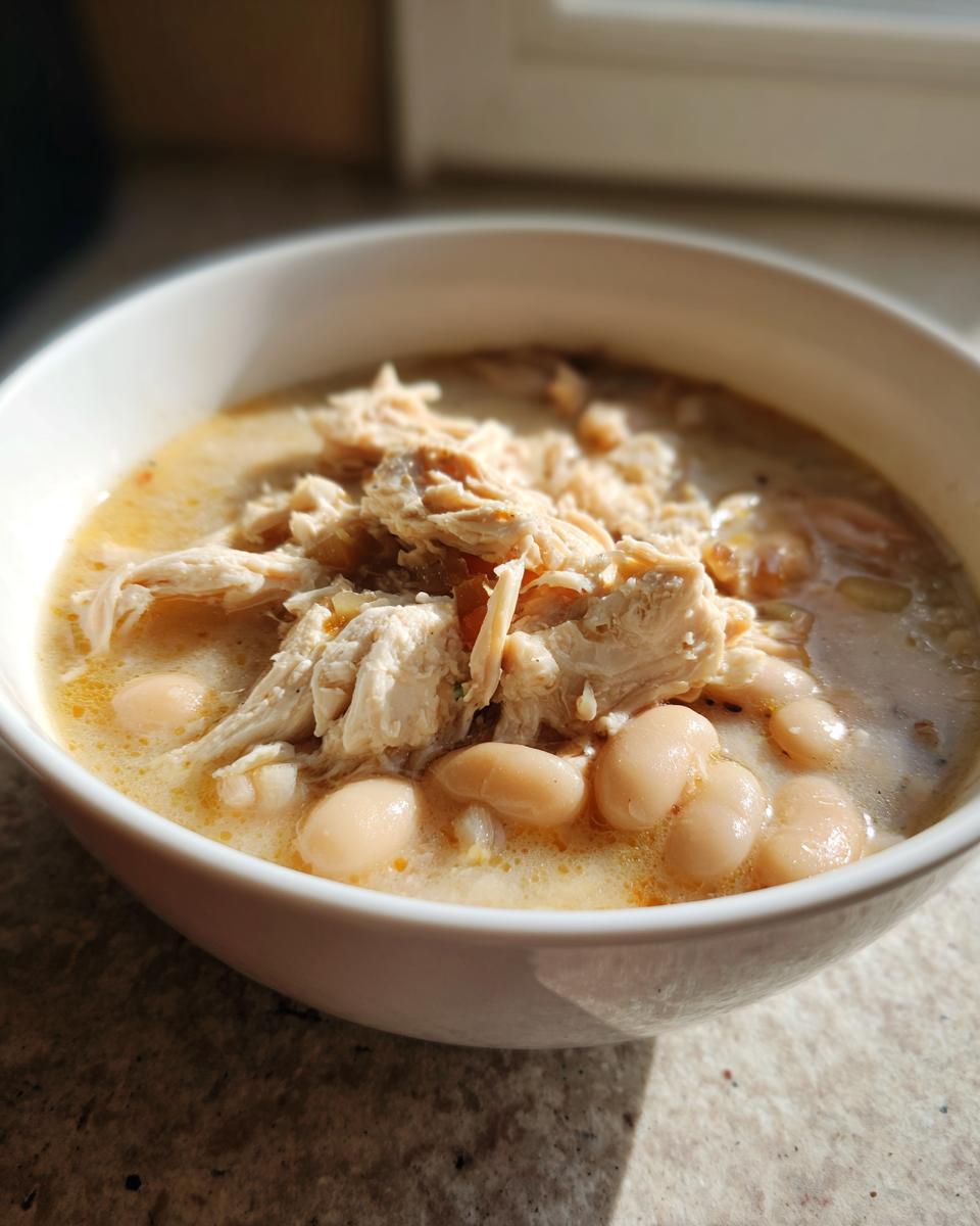 Close-up of a white bowl filled with creamy white chicken chili, topped with shredded white chicken and cannellini beans.