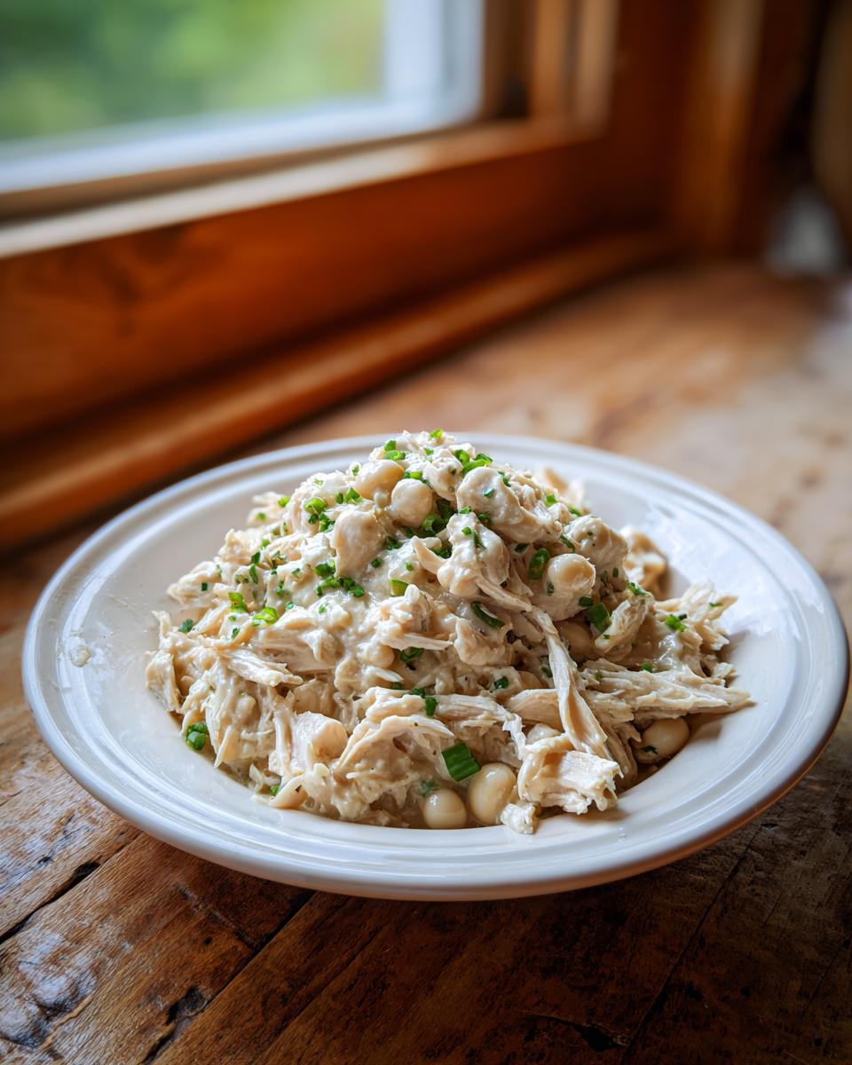 A bowl of creamy white chicken chili with beans and chives served on a rustic wooden table near a window.