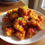 A plate piled high with crispy, glazed pieces of orange chicken recipe, garnished with sesame seeds and green onions.