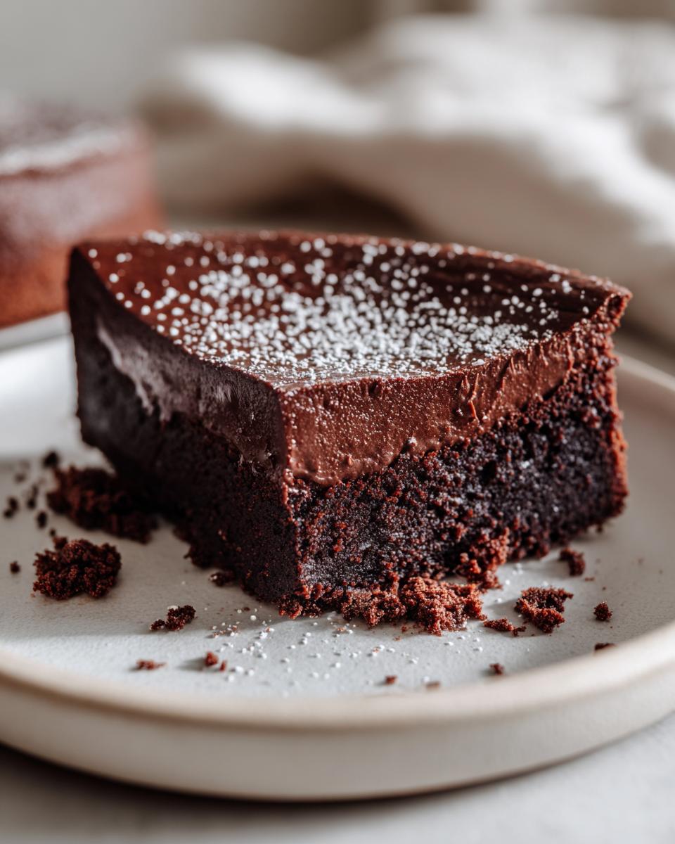 Close-up of a dense slice of flourless chocolate cake topped with ganache and dusted with powdered sugar.