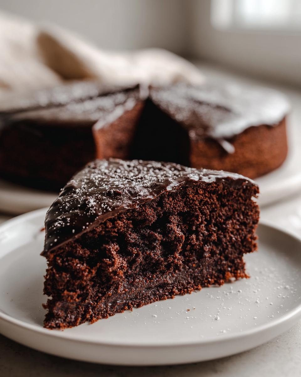 A close-up of a moist slice of flourless chocolate cake topped with dark chocolate glaze and powdered sugar.