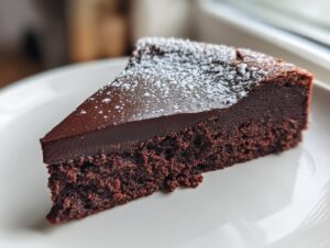 A close-up of a decadent slice of flourless chocolate cake, topped with a smooth ganache layer and dusted with powdered sugar.