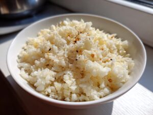 Close-up of a white bowl filled with fluffy, seasoned cauliflower rice, illuminated by natural light.