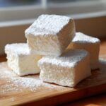 Four fluffy cubes of homemade marshmallows generously dusted with white powdered sugar, stacked on a wooden board.