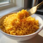 A golden bowl of fluffy turmeric rice being lifted slightly by a gold fork near a bright window.