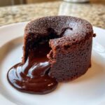 Close-up of a perfectly baked chocolate lava cake with molten chocolate oozing out onto a white plate.