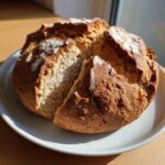 A rustic, round loaf of freshly baked soda bread sitting on a white plate, with a wedge cut out showing the soft interior crumb.