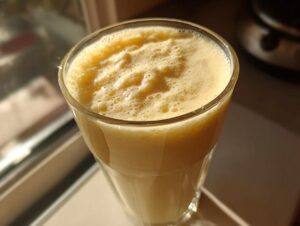Close-up of a tall glass filled with a thick, frothy yellow banana smoothie, catching warm sunlight near a window.