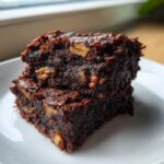 Two fudgy date brownies stacked on a white plate, showing a rich, dark chocolate texture with visible chunks of dates.