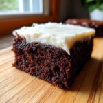 Close-up of a rich, dark slice of gingerbread brownies topped with thick white frosting on a wooden board.