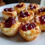 Close-up of golden baked brie bites made with puff pastry, melted brie, and a dollop of bright red jam.