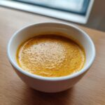 A close-up of rich, golden-orange beer cheese dip with a slight froth, served in a small white bowl on a wooden surface.