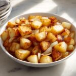 A white bowl filled with perfectly golden brown fried potatoes mixed with sautéed onions.