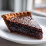 Close-up of a rich, dark slice of chocolate chess pie with a flaky crust on a white plate.