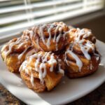 A close-up of several gooey cinnamon roll bites drizzled with white icing, sitting on a white plate near a window.