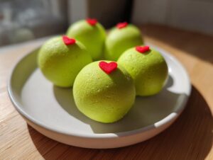 Five bright green grinch oreo balls, each topped with a tiny red heart, arranged on a light gray plate.