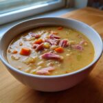 A close-up of a white bowl filled with thick, creamy navy bean soup topped with shredded ham and diced carrots.