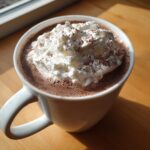 Close-up of a mug of rich homemade hot cocoa topped with a generous swirl of whipped cream and cocoa powder.