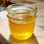 Close-up of a bright yellow homemade salad dressing, likely a vinaigrette, settling in a small glass jar.