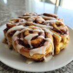 Close-up of several freshly baked christmas tree cinnamon rolls generously drizzled with white icing on a white plate.