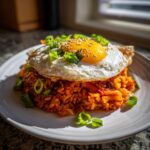 A mound of vibrant red kimchi fried rice topped with a sunny-side-up egg, sesame seeds, and sliced green onions.