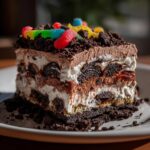 Close-up of a decadent slice of four-layer dirt cake featuring cream, chocolate pudding, and Oreo pieces, topped with gummy worms.