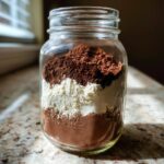 A glass mason jar filled with layered ingredients for hot cocoa mix, featuring dark and light brown powders.