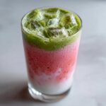 Close-up of a tall glass containing a layered strawberry matcha drink with milk and ice cubes on top.