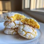 A stack of bright yellow lemon crinkle cookies dusted heavily with powdered sugar, one cookie broken open to show the soft interior.