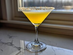 A vibrant yellow lemon drop martini in a chilled coupe glass with a sparkling sugar rim, set against a bright window.
