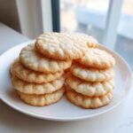 A stack of freshly baked, light golden sugar cookies with a textured top, resting on a white plate near a window.