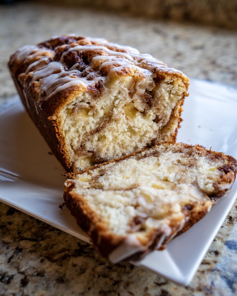 A loaf of moist apple bread, partially sliced, showing a cinnamon swirl and drizzled with white icing.