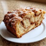 A thick slice of moist apple bread featuring a cinnamon swirl and topped with a sweet vanilla glaze.
