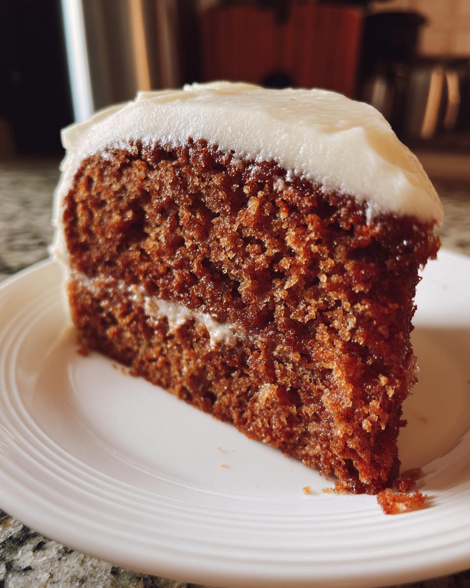 Close-up of a moist slice of dark brown applesauce cake topped with thick white frosting on a white plate.