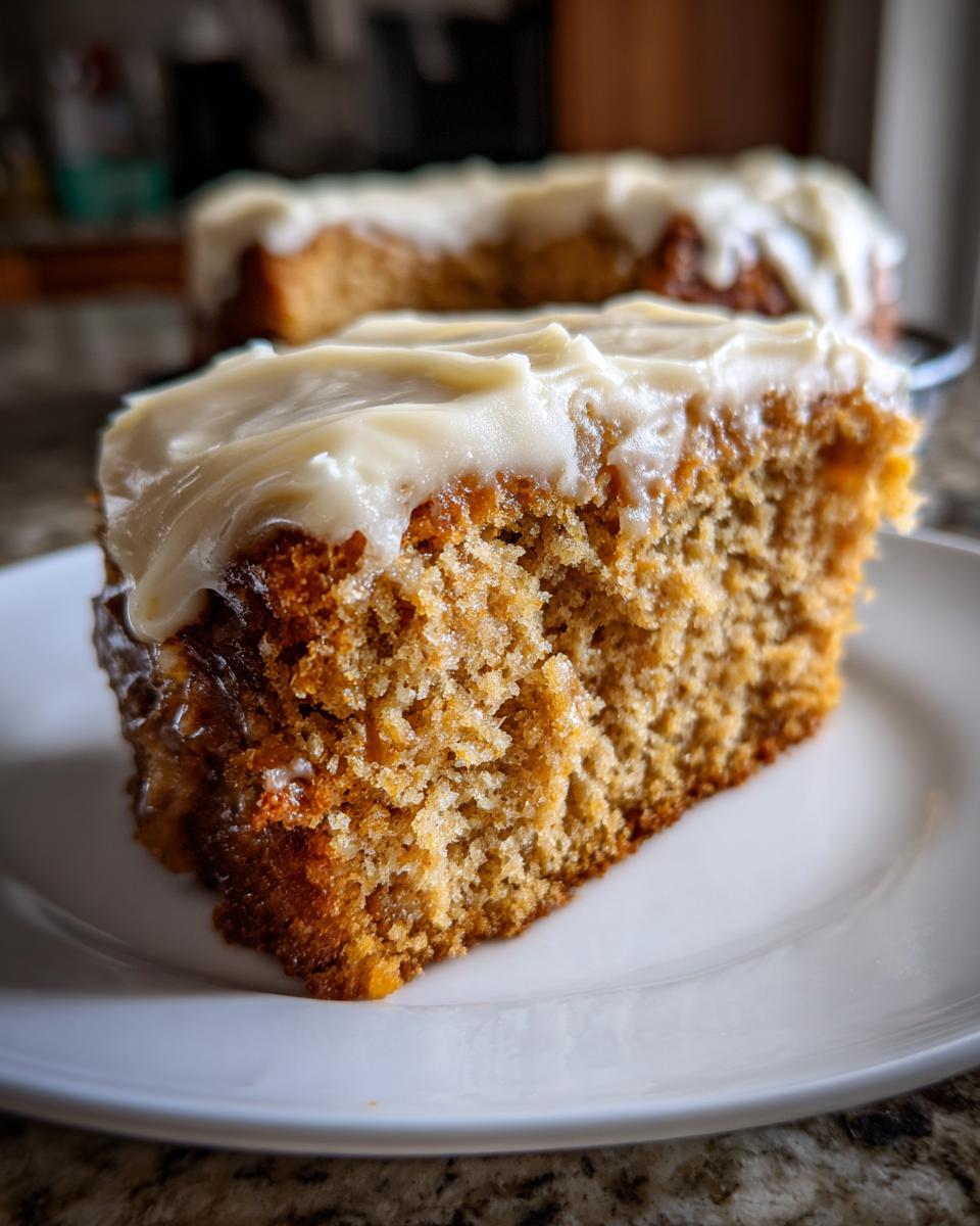 Close-up of a moist slice of applesauce cake topped with thick, creamy white frosting on a white plate.