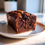 A close-up of a freshly baked, moist chocolate bread loaf studded with dark chocolate chips, resting on a light plate.