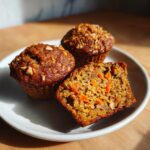 Three moist morning glory muffins on a white plate, one cut in half showing shredded carrots inside.