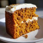 Close-up of a moist, orange-hued slice of spice cake layered with white frosting, sitting on a white plate.